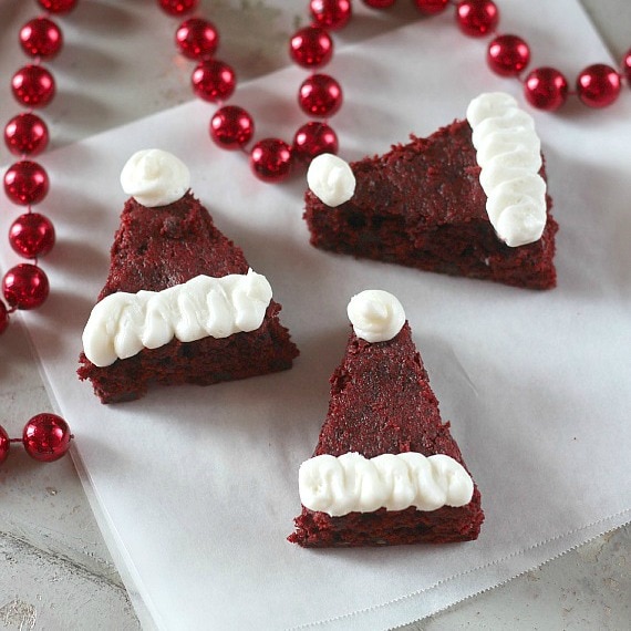 Fun and festive Santa hat brownies are easy to make from a box of red velvet cake mix and fluffy white frosting. Perfect for the holidays! Red velvet Santa hat brownies decorated with white frosting next to a string of red beads.