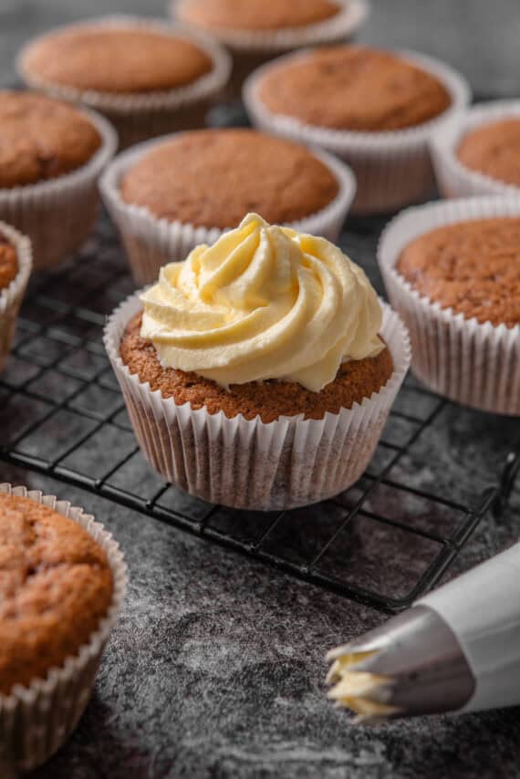 A gingerbread cupcake topped with a frosting swirl on a wire rack, with unfrosted cupcakes in the background.