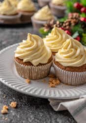 Three frosted gingerbread cupcakes on a white plate with Christmas decor and more cupcakes in the background.