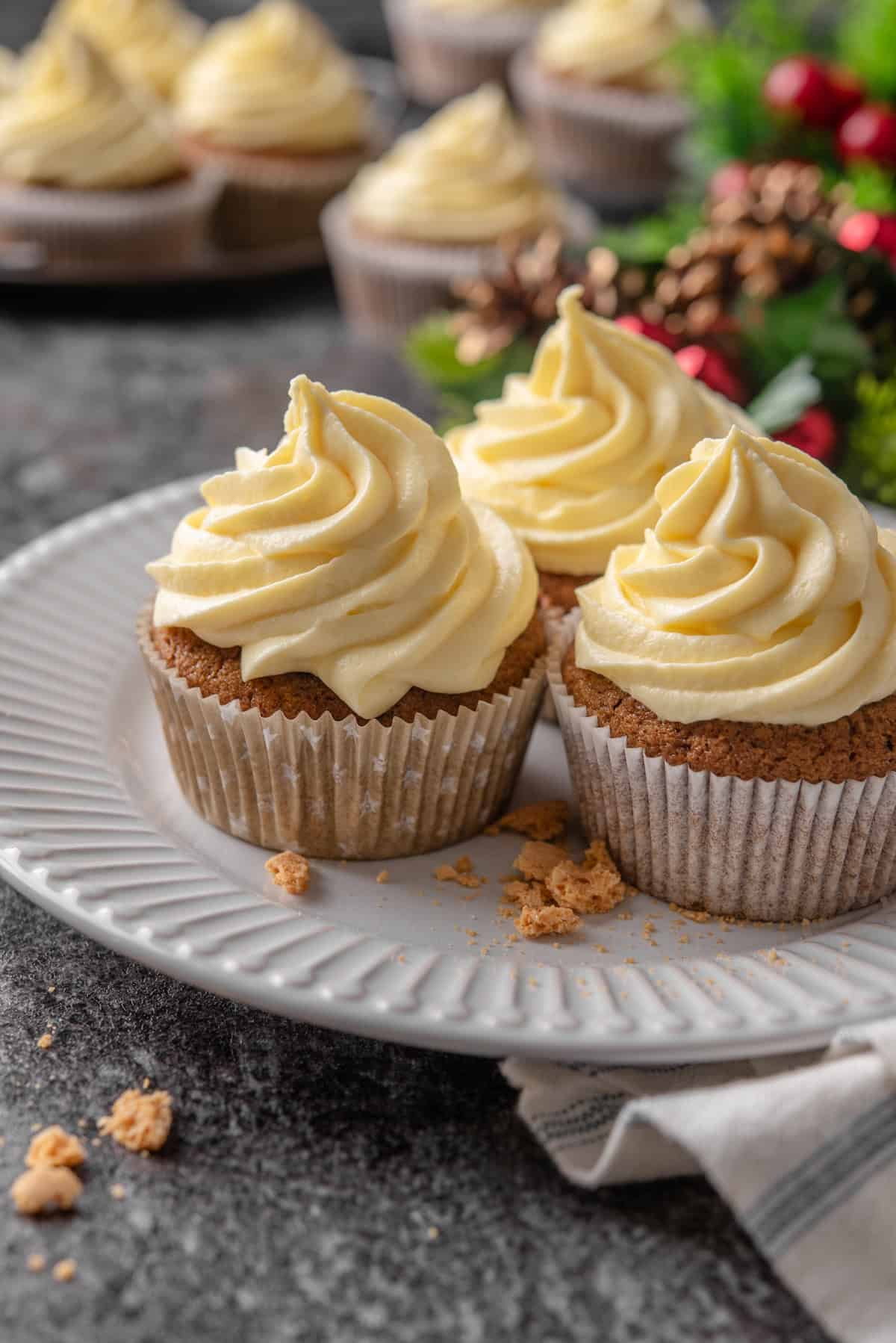 Three frosted gingerbread cupcakes on a white plate with Christmas decor and more cupcakes in the background.