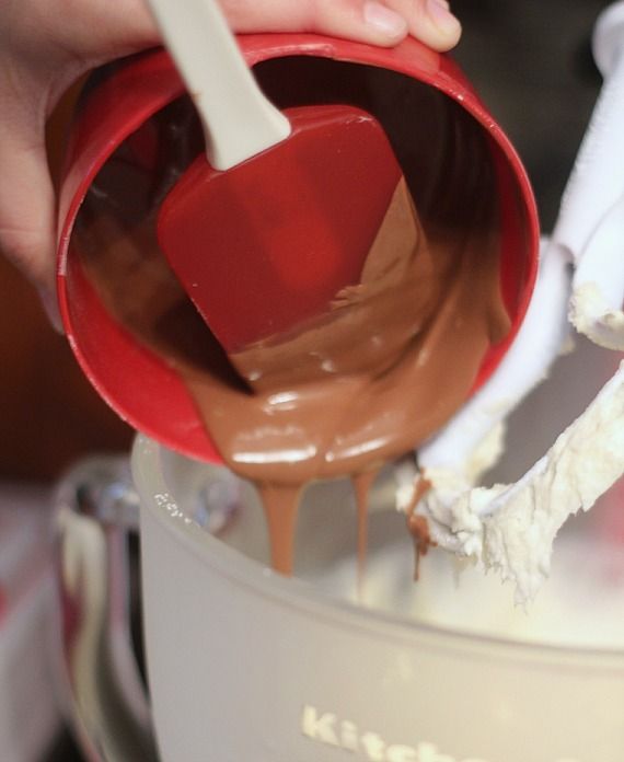 A measuring cup of melted chocolate being poured into a stand mixer bowl