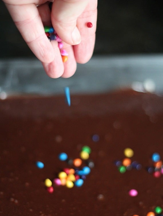 Rainbow sprinkles being spread on top of brownie batter in a baking pan