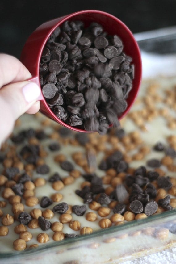 Chocolate chips being poured from a measuring cup into a baking dish of sweetened condensed milk and caramel bits