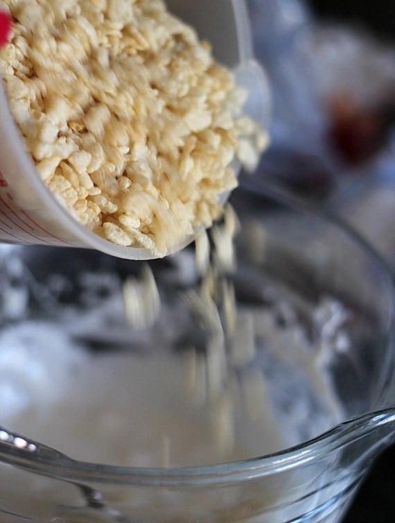 A cup of rice krispies being poured into a bowl of melted marshmallows