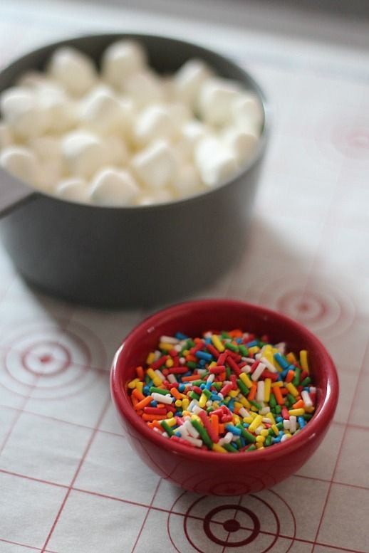 A measuring cup of mini marshmallows and a small bowl of rainbow sprinkles