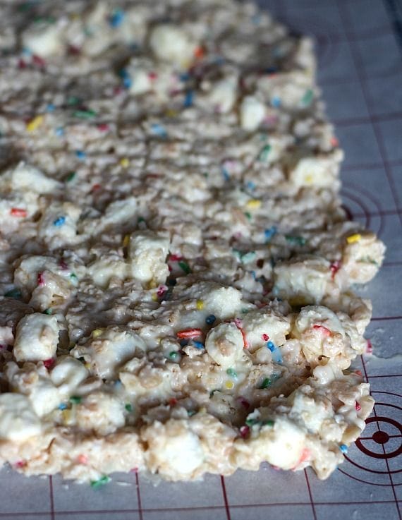 Rice krispie treat batter with rainbow sprinkles spread on a parchment-lined baking sheet