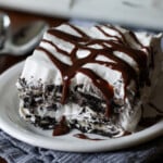 A layered whipped cream and Oreo icebox cake dessert slice on a plate topped with chocolate sauce