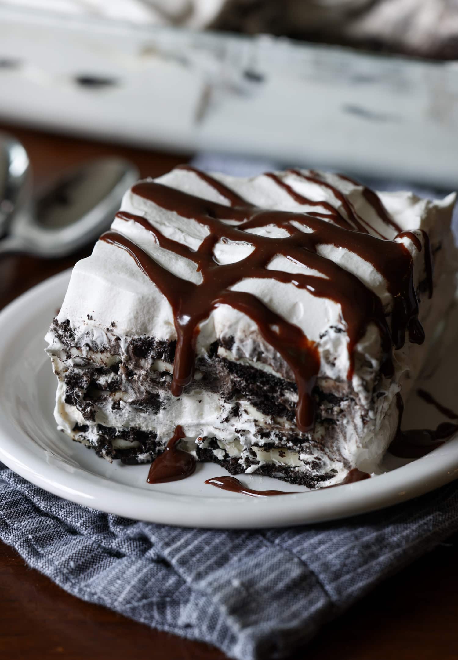 A layered whipped cream and Oreo icebox cake dessert slice on a plate topped with chocolate sauce