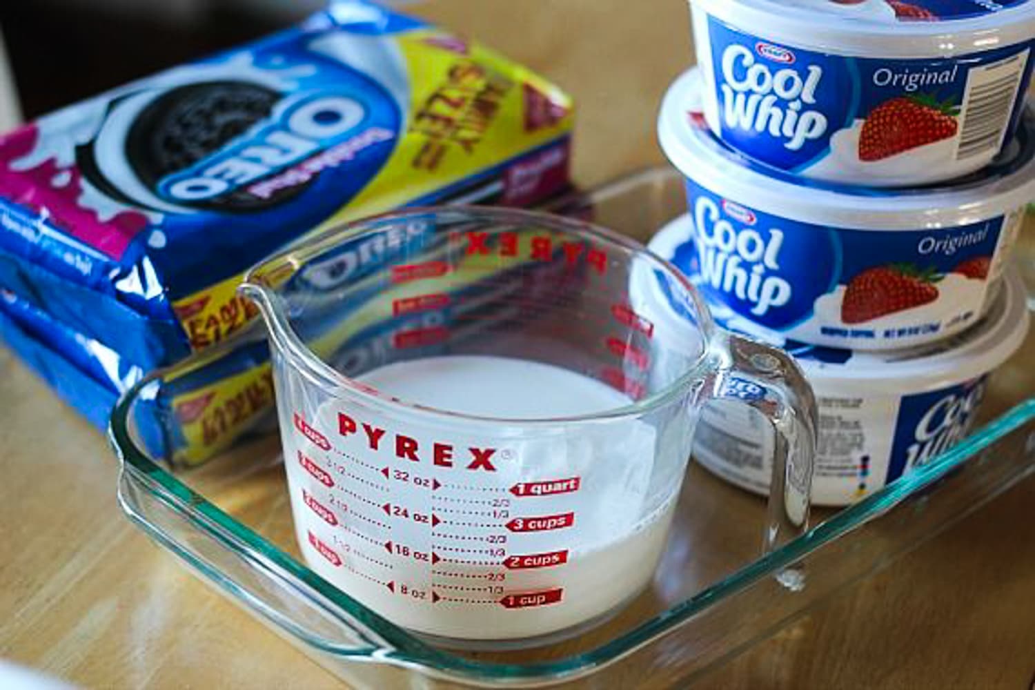 milk, oreo cookies, and three tubs of cool whip on the counter with a 9x13 baking dish