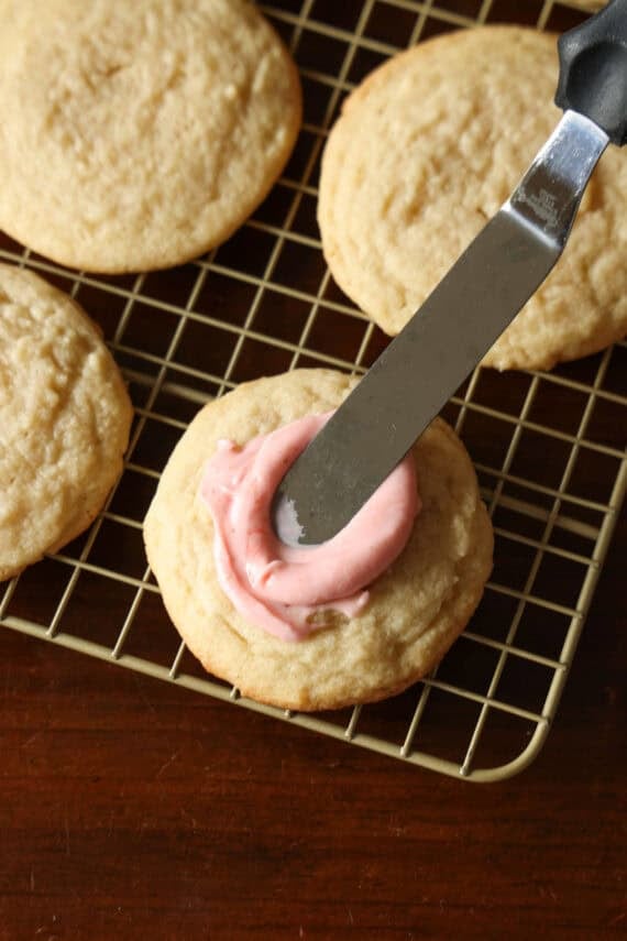 Using an offset spatula to spread pink strawberry frosting onto a sugar cookie.