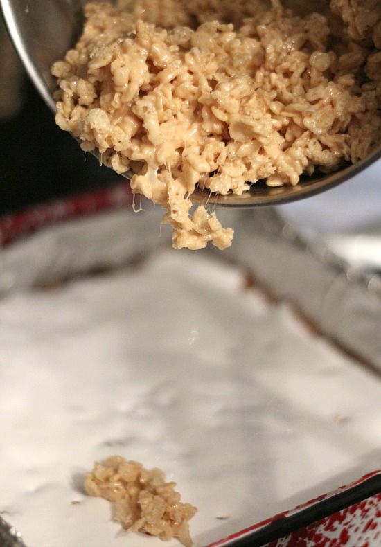 Caramel rice krispie treat mixture being added to a pan of peanut butter blondies with marshmallow topping