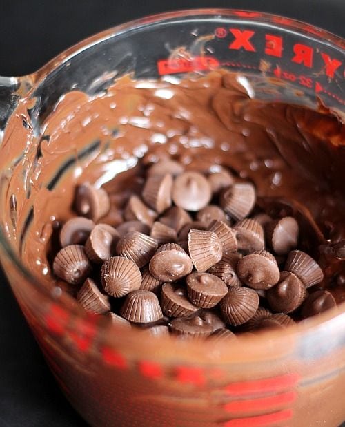 Mini peanut butter cups in a glass measuring cup of melted chocolate and chopped peanuts