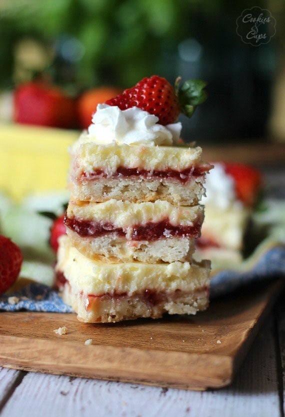 A stack of three Strawberry Lemon Cheesecake Bars topped with whipped cream and a strawberry on a wooden board.