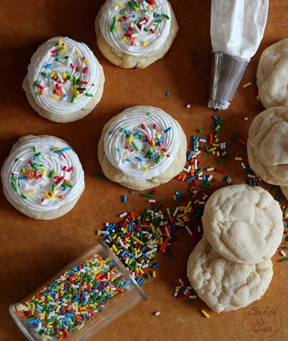 Soft sugar cookies topped with buttercream and sprinkles on a counter, next to unfrosted cookies and sprinkles spilling out of the container