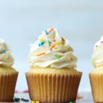 Three fluffy, soft vanilla cupcakes topped with swirls of vanilla frosting and sprinkles in a row.