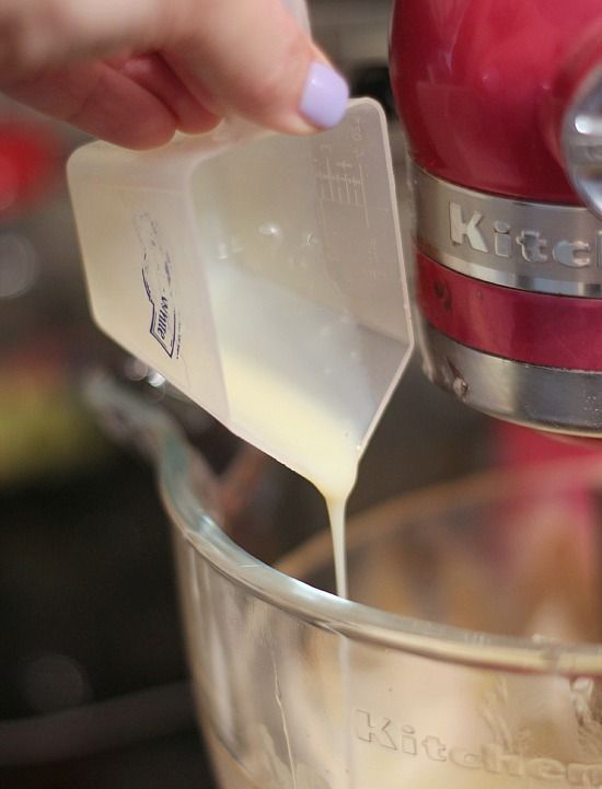 A measuring cup of sweetened condensed milk being poured into the bowl of a stand mixer