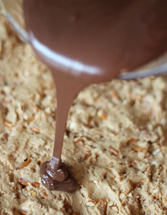 Melted chocolate peanut butter mixture being poured over peanut butter pretzel dough in a pan