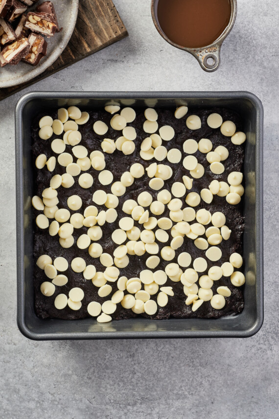 White chocolate chips are added on top of the chocolate batter layer in a baking pan.
