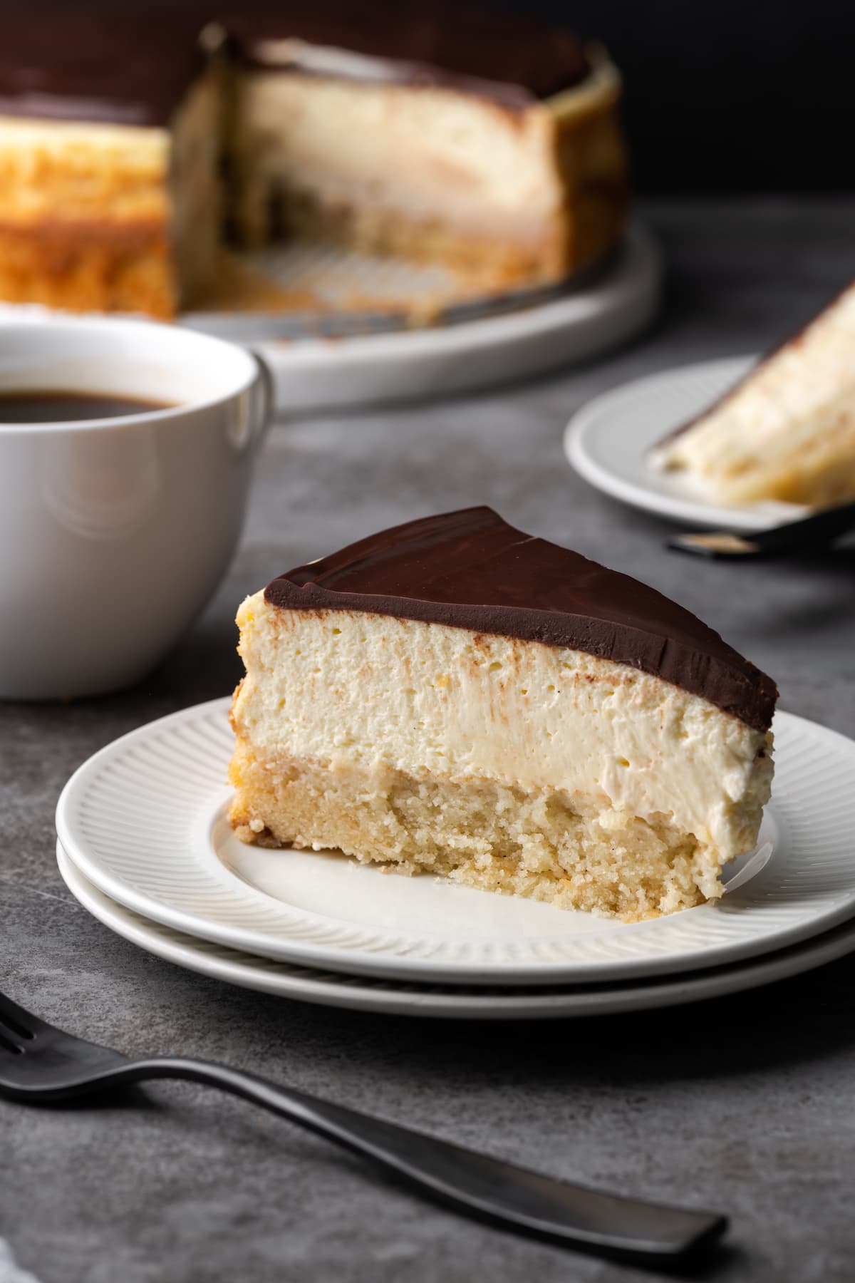 A slice of Boston Cream Pie cheesecake on a plate, with a coffee mug and the remaining cake in the background.