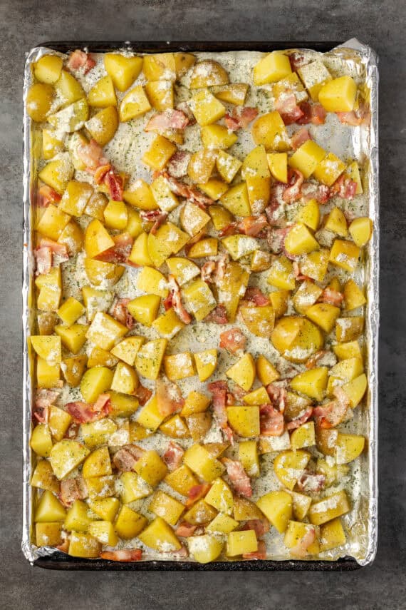 Sliced ​​potatoes and bacon topped with ranch seasoning on a lined baking sheet.