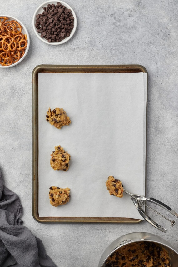 A cookie scoop is used to place balls of pretzel chocolate chip cookie dough onto a lined baking sheet.