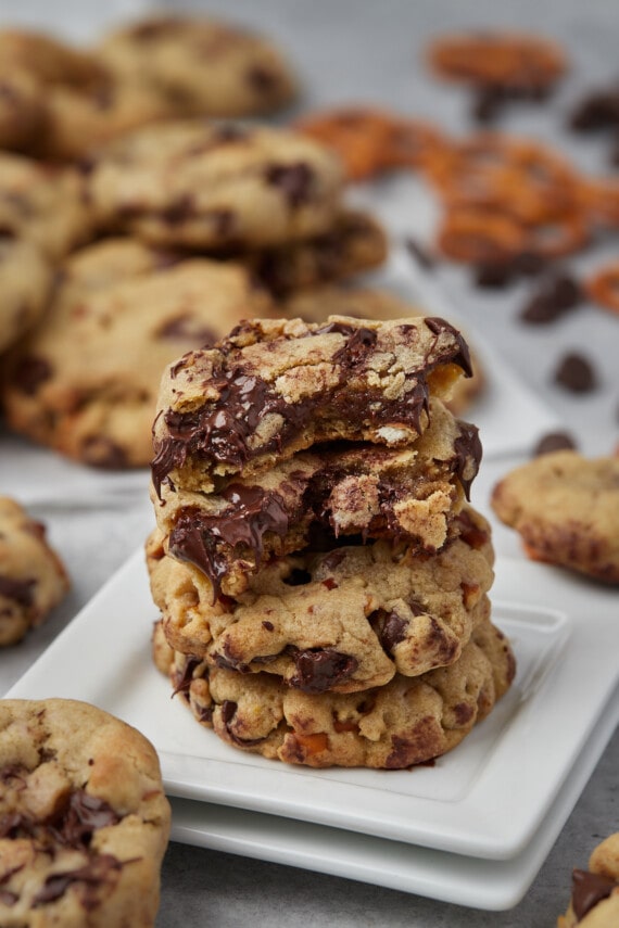 Salty pretzel chocolate chip cookies stacked on a square white plate, surrounded by cookies.