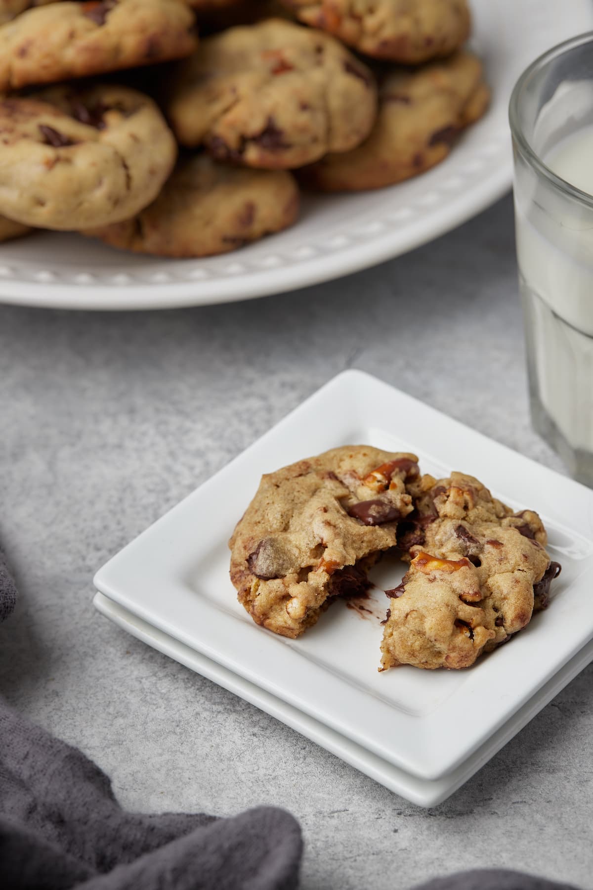 Two halves of a pretzel chocolate chip cookie on a plate next to a glass of milk and a plate full of cookies.