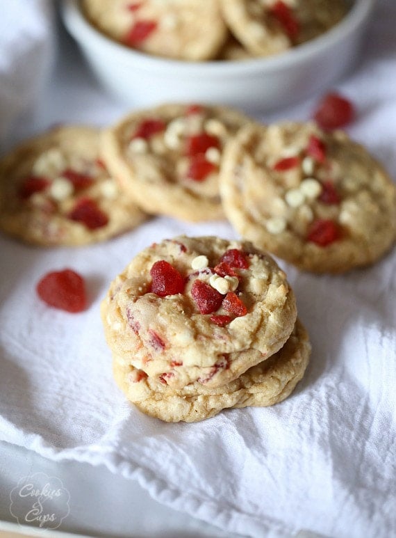 Assorted strawberry white chocolate chip cookies scattered over a white table cloth, with more cookies in a white bowl in the background.