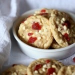 Strawberry white chocolate chip cookies in a white bowl, with more cookies scattered around on a white table cloth.
