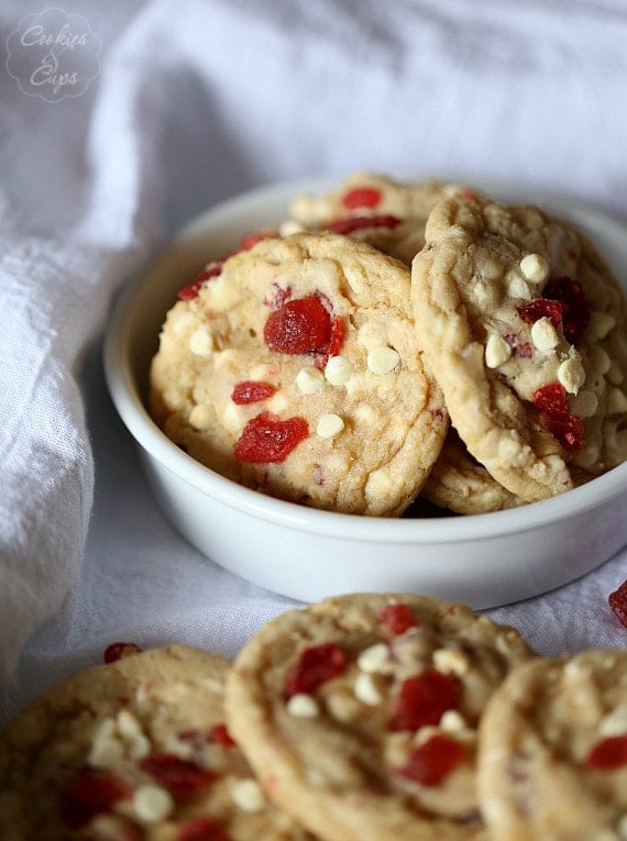 Strawberry white chocolate chip cookies in a white bowl, with more cookies scattered around on a white table cloth.