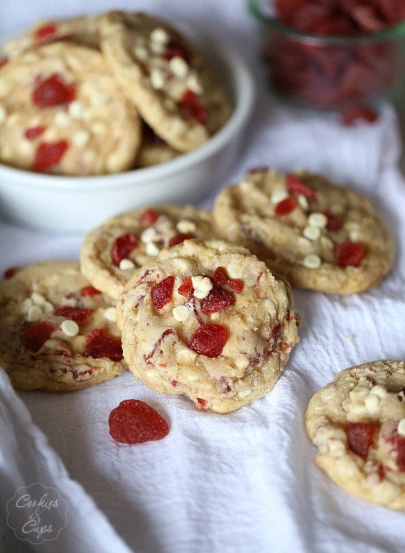 Assorted strawberry white chocolate chip cookies scattered over a white table cloth, next to more cookies in a white bowl.