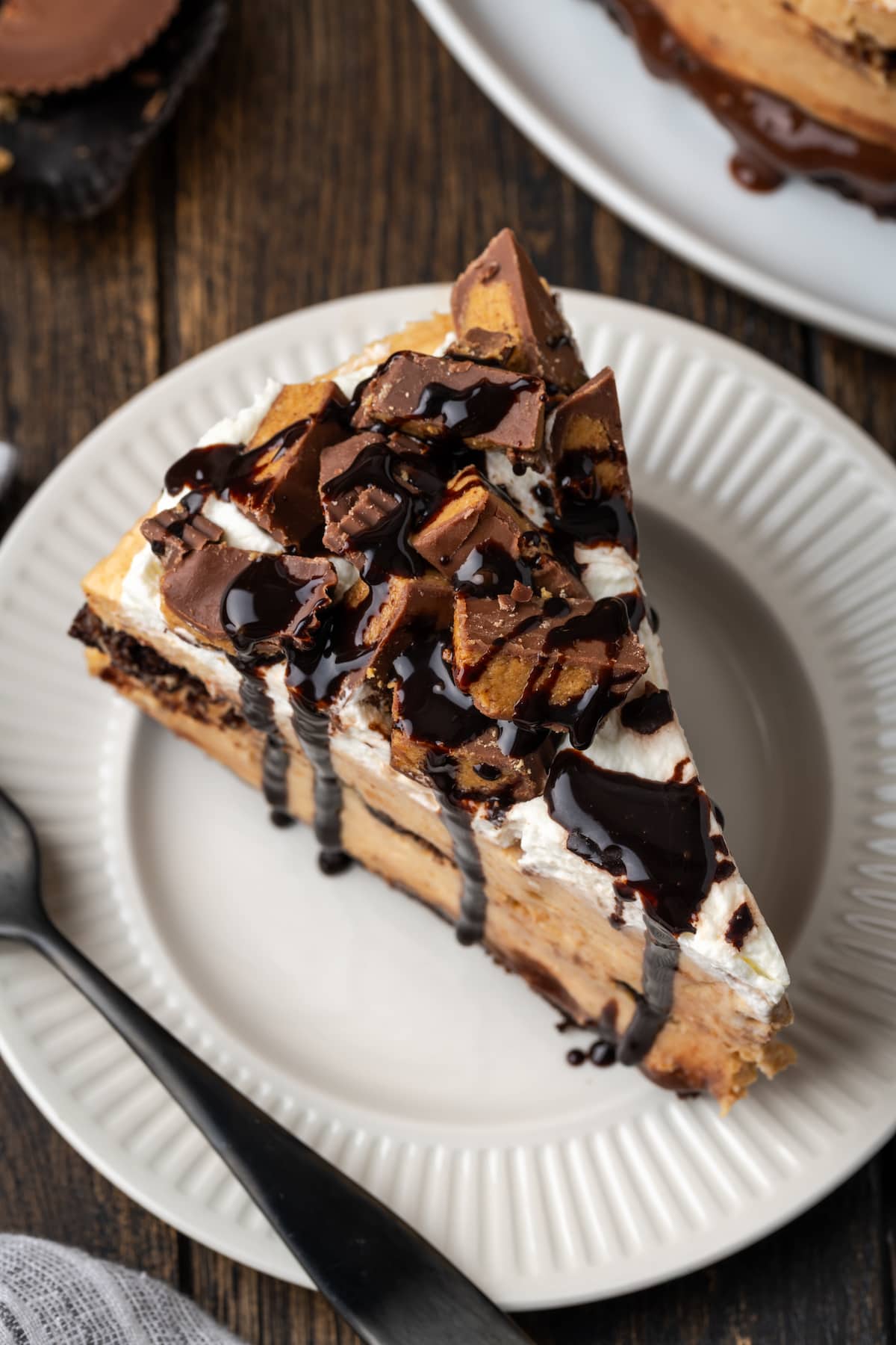 A slice of brownie cheesecake garnished with peanut butter cups and fudge sauce on a white plate next to a fork.