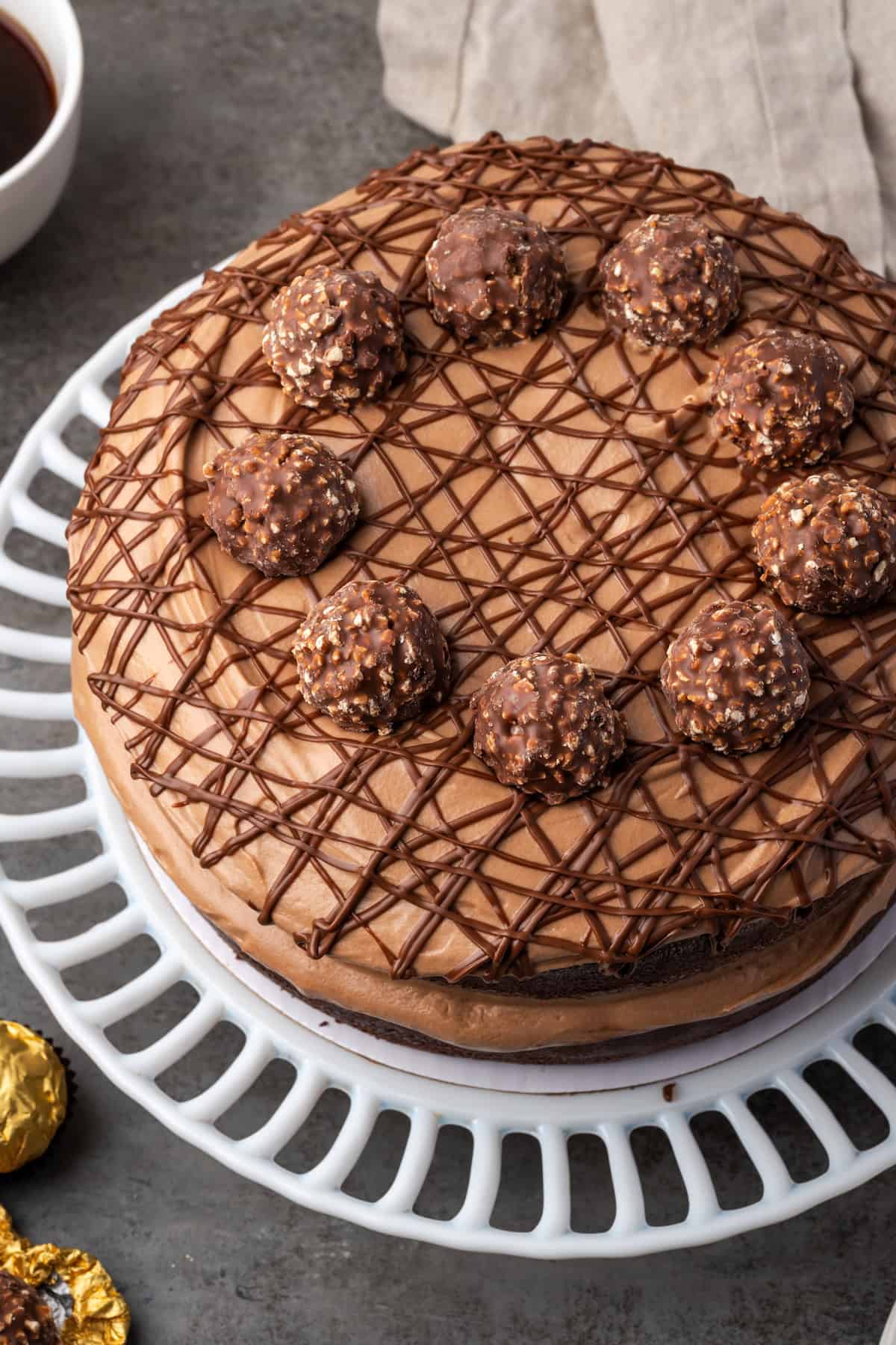 This chocolate hazelnut cake has thick, fudgy chocolate cake layers filled with creamy chocolate hazelnut spread and silky cheesecake mousse. Overhead view of chocolate hazelnut cake drizzled with chocolate and topped with chocolate hazelnut candy on a cake stand.