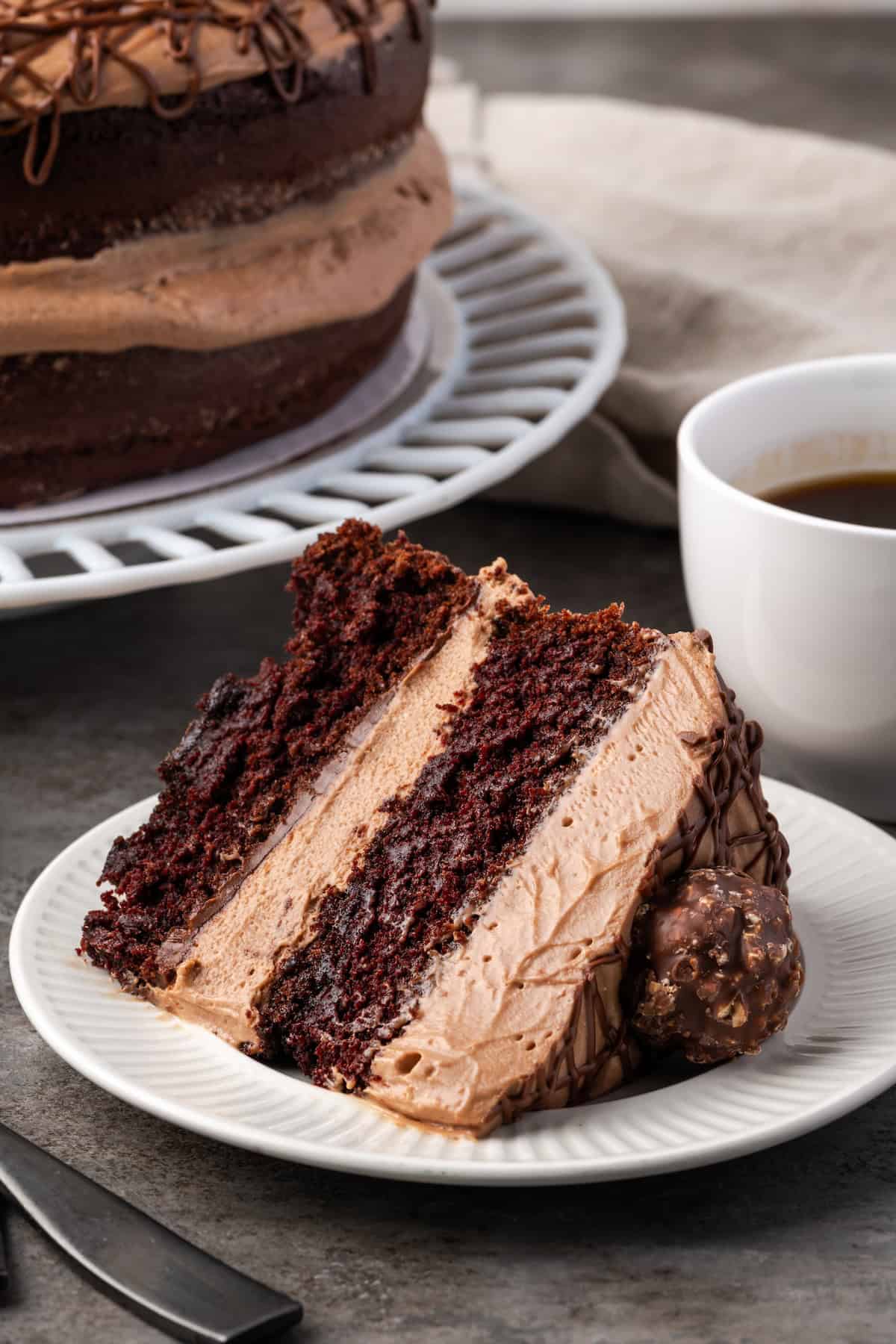 A slice of chocolate hazelnut cake on a white plate with a cup of coffee and the rest of the cake in the background.