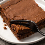 A fork cuts into a slice of chocolate sheet cake on a white plate.