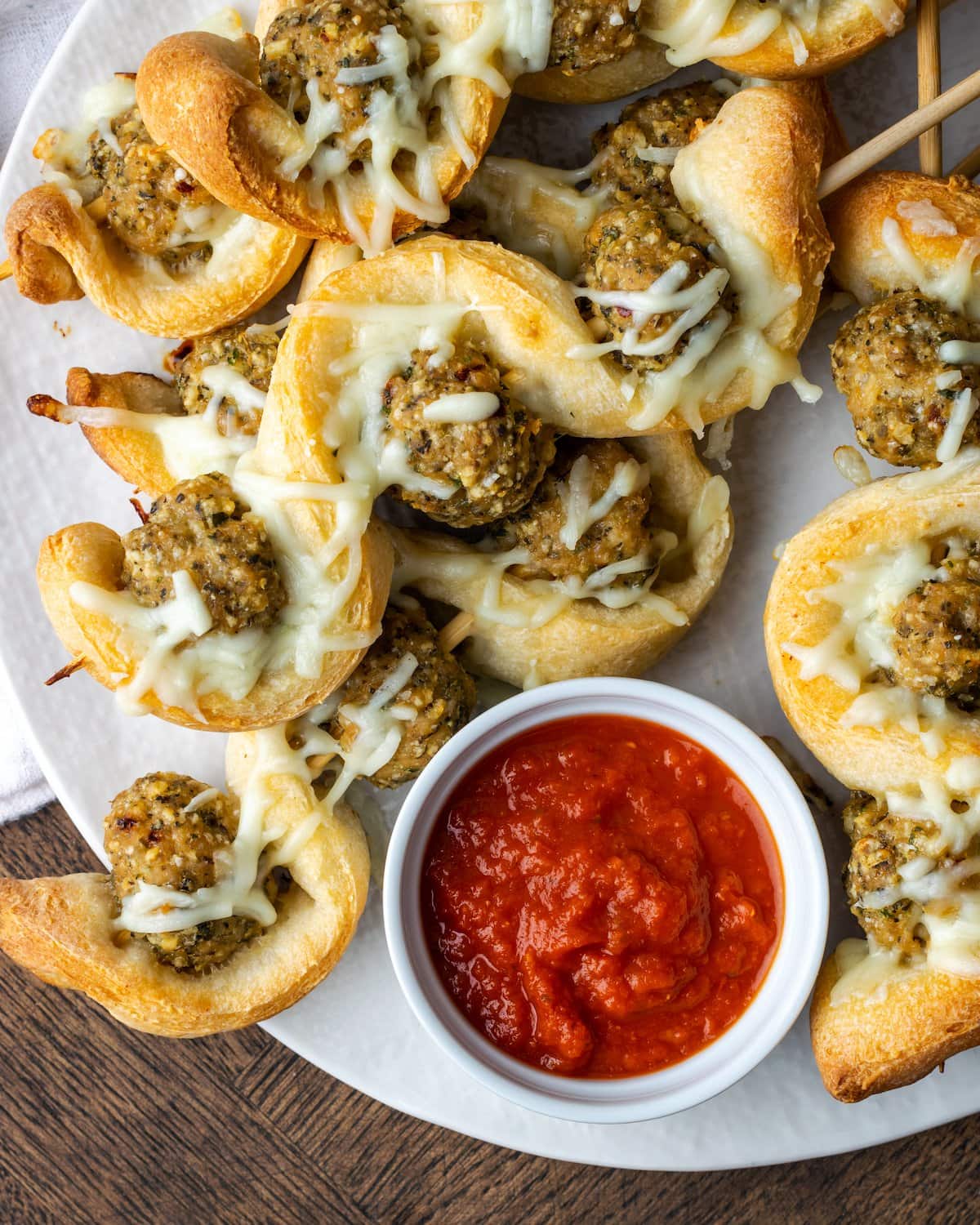 Top view of a plate of meatball subs on a stick with a bowl of marinara sauce.