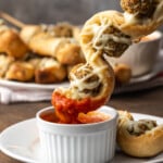 A meatball sub on a stick is dipped into a ramekin of marinara sauce.