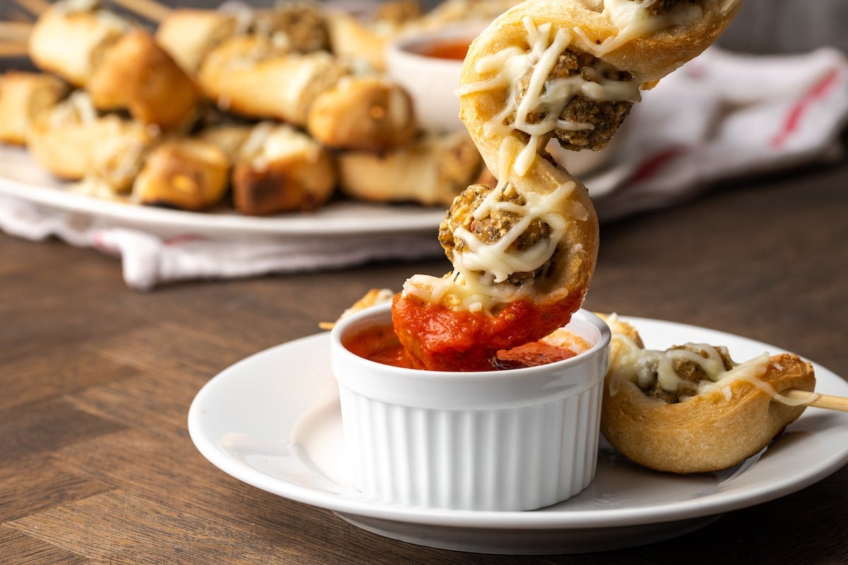 A meatball sub on a stick is dipped into a ramekin of marinara sauce.