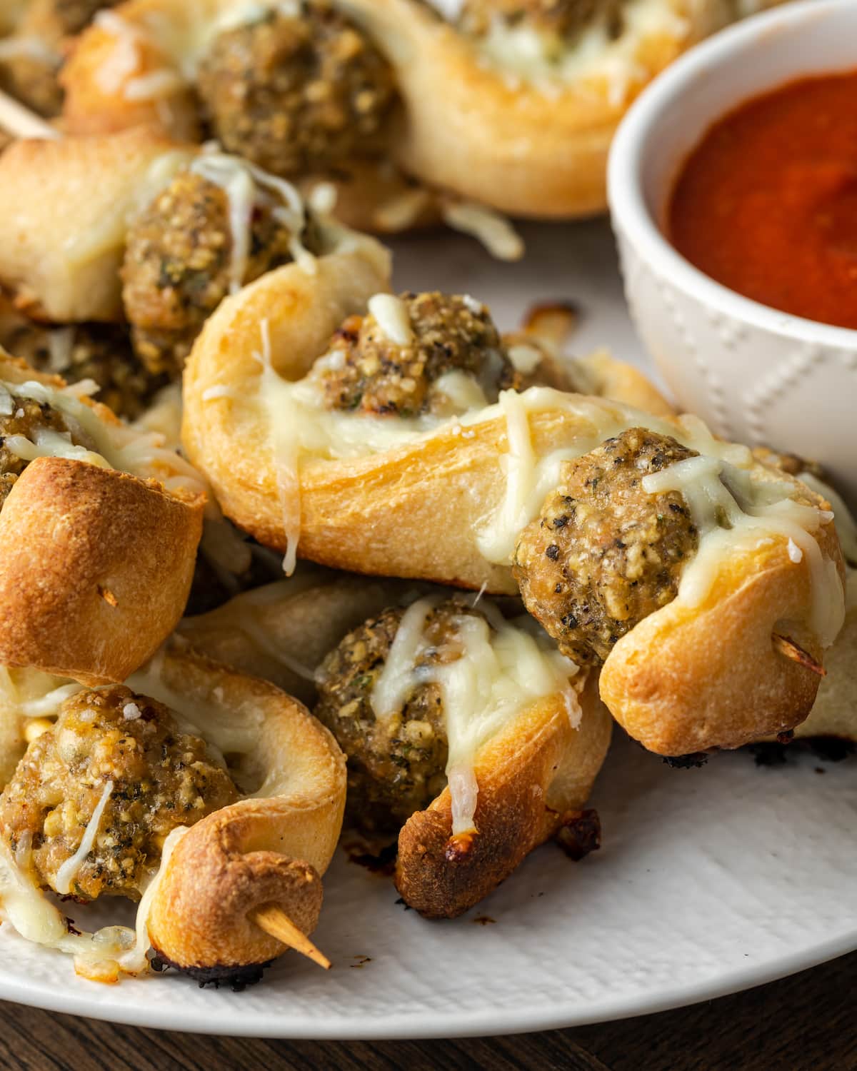 A plate of meatball subs on a stick with a bowl of marinara sauce.