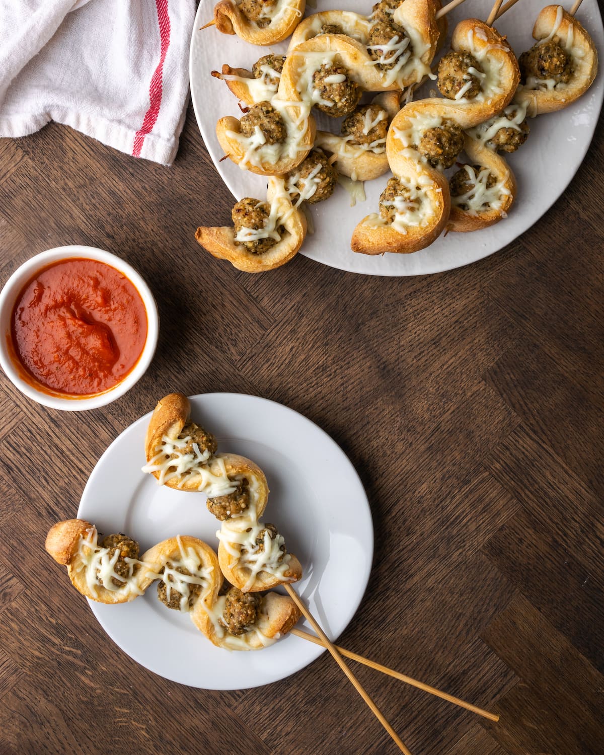 Top view of two baked meatball subs on sticks on a white plate with a bowl of marinara sauce, next to a full platter of meatball subs.