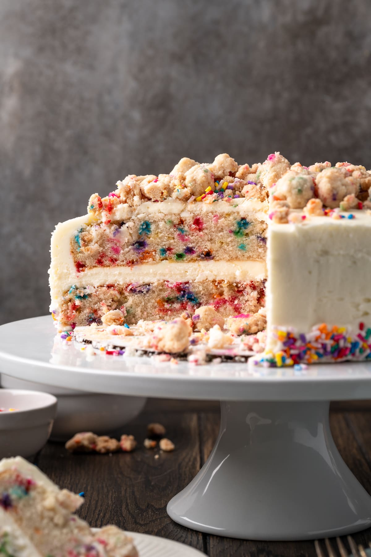 Frosted confetti cake on a cake stand topped with funfetti streusel and sprinkles, with a large slice missing.