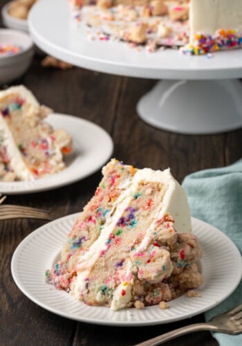 A slice of frosted confetti cake on a white plate, with a second slice on a plate next to a cake stand in the background.