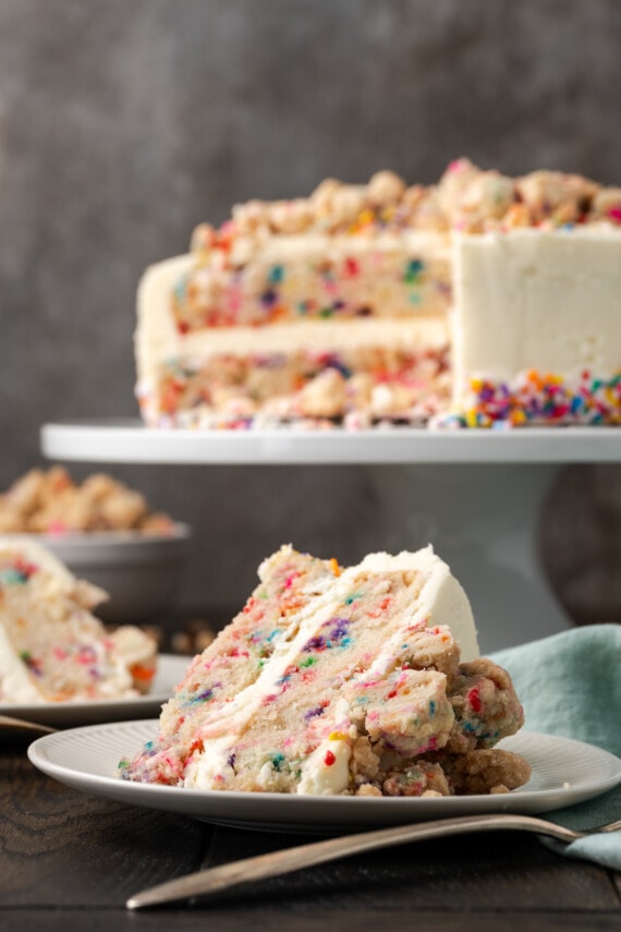Two slices of confetti cake on plates with the rest of the cake on a cake stand in the background.