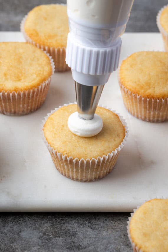 A piping tip piping frosting onto an almond cupcake surrounded by more unfrosted cupcakes.