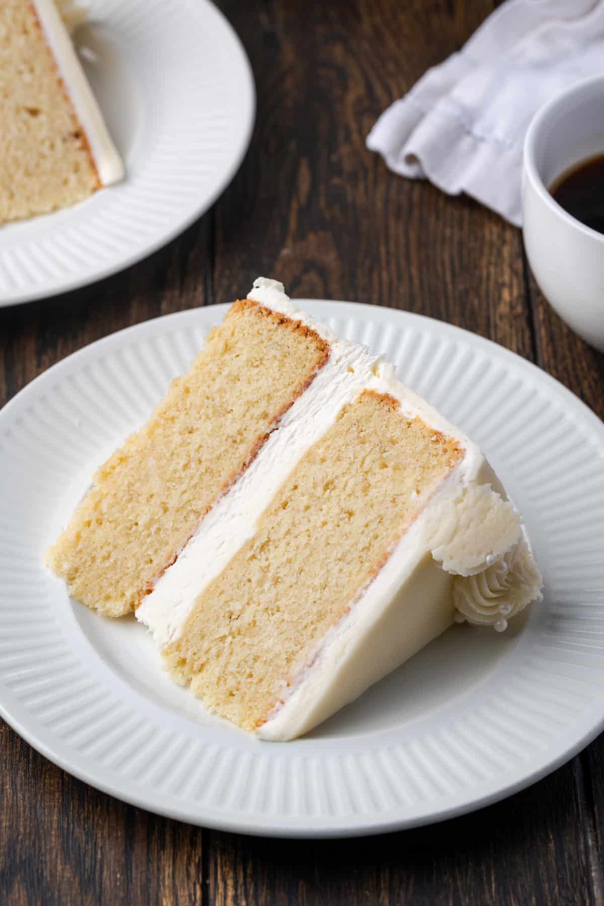 A slice of vanilla cake on a white plate, with a second slice in the background.