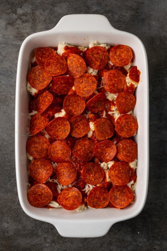 A final layer of pepperoni is added to pizza casserole in a baking dish.