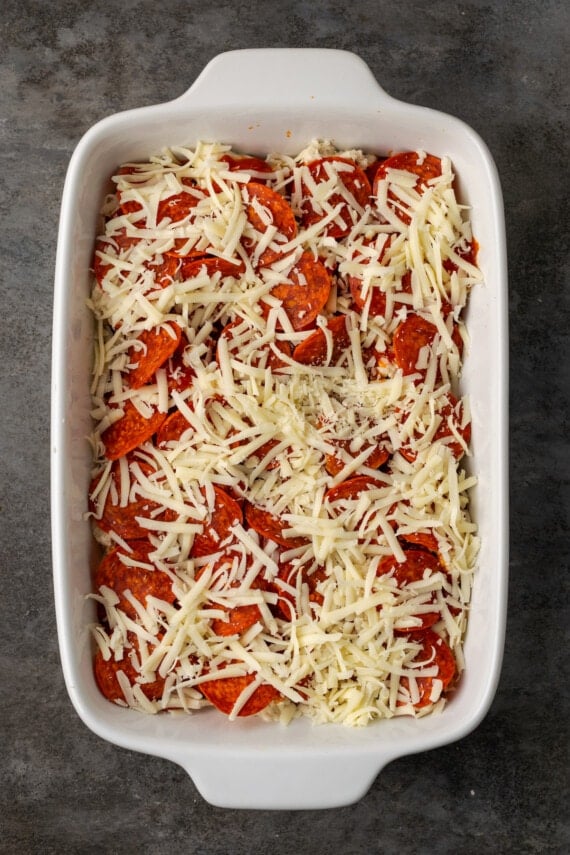 A final layer of shredded cheese is added to pizza casserole in a baking dish.