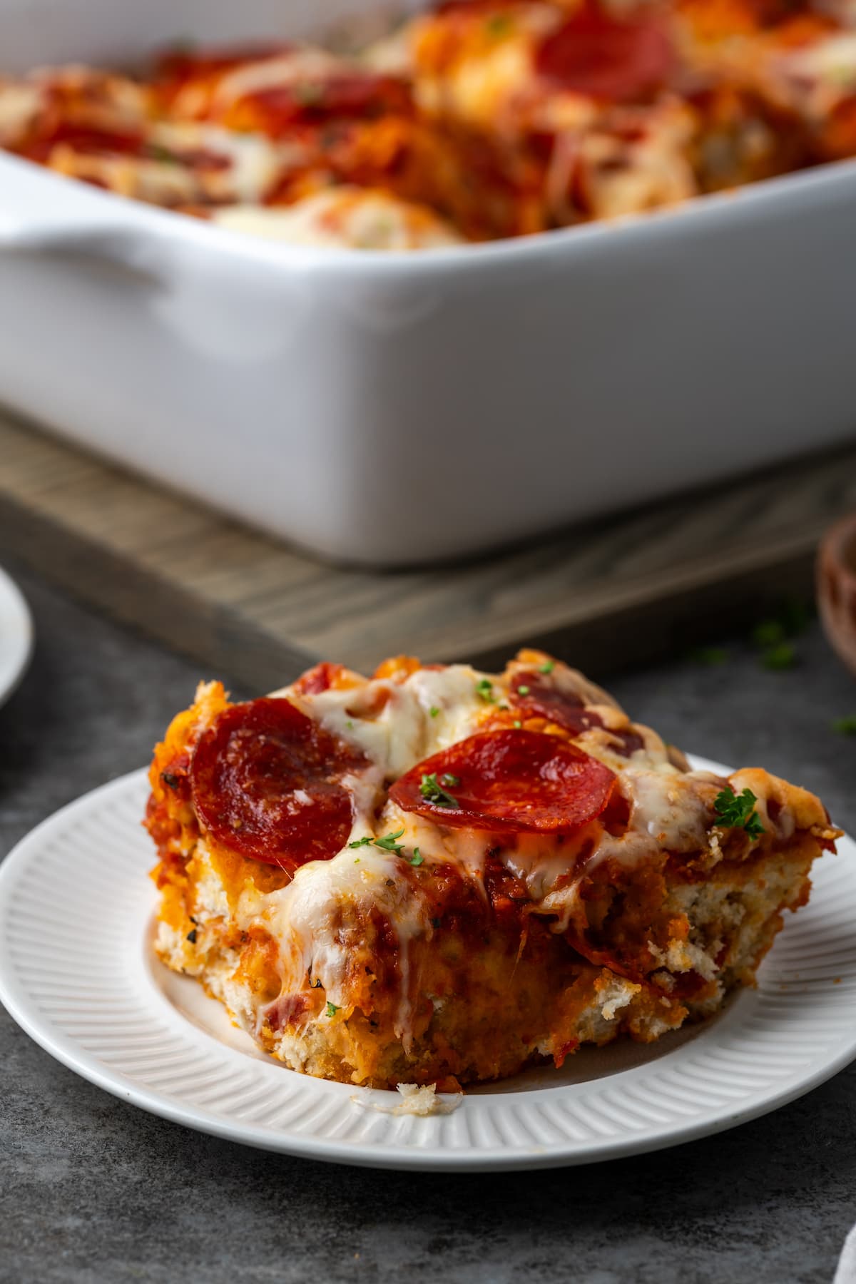A serving of pizza casserole on a white plate, with the rest of the casserole in a baking dish in the background.