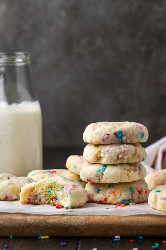 Side view of a stack of funfetti cookies on a parchment-lined wooden cutting board, next to one cookie with a bite missing.