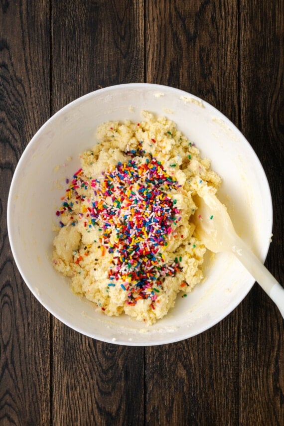 Sprinkles added into Funfetti cake mix cookie dough in a white mixing bowl with a spatula.